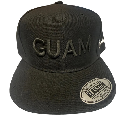 A black snapback cap with 'GUAM' embroidered in black on the front, featuring a white oval 'Klassick' sticker on the brim and a white script logo on the side, displayed on a light surface with a colorful background.