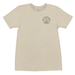 A cream-colored short-sleeve t-shirt with a circular graphic on the left chest, laid flat on a gray wood plank floor. A person's feet wearing black flip-flops are visible at the bottom edge of the frame.