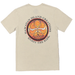 A cream-colored short-sleeve t-shirt laid flat on a gray wood floor, featuring a circular graphic on the back with the text 'HAWAIIAN ISLAND CREATIONS' and 'ENJOY THE RIDE' around an orange and yellow sun-and-mountain design. Parts of a person's feet in black flip-flops are visible at the bottom corners.